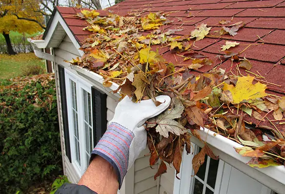 Roofer performing roofing maintenance, clearing overfilled gutters from a residential home with red shingles.