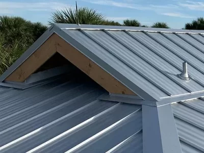 A newly installed gray corrugated metal roof with a triangular gable vent covered by an angled roof flashing. A metal pipe protrudes from the right side, surrounded by a backdrop of green trees and blue sky.