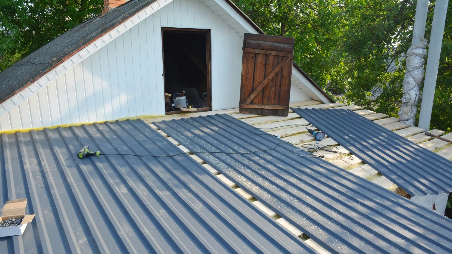 A partially renovated roof on an old house with gray corrugated metal sheets partially installed on a wooden frame. An open attic door with visible renovation tools inside is to the right, and several tools and screws are visible on the metal sheets.